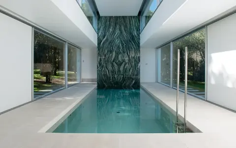 Piscina interior con pared de mármol verde veteado, rodeada de ventanales que dan a un jardín.