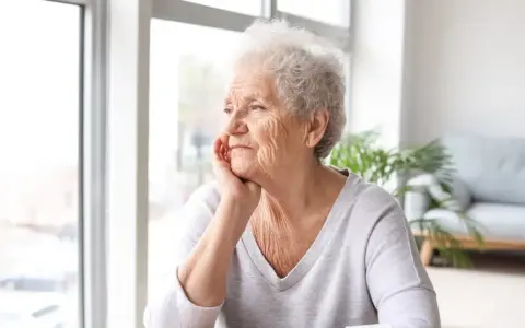 Anziana con mano sul mento, sguardo pensieroso verso la finestra. Potrebbe riflettere sulla pseudodemenza depressiva.