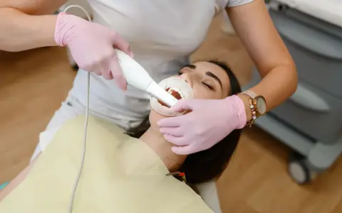 Dentystka w różowych rękawiczkach wykonuje zabieg piaskowania zębów. Czy piaskowanie zębów boli? Pacjentka ma otwarte usta i rozwierak.