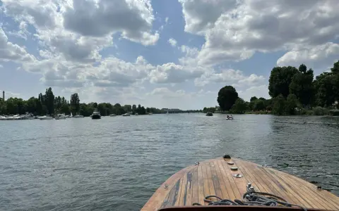Blick vom Bug eines Holzbootes auf einen Fluss mit vielen Booten und grünen Ufern. Ein schöner Tag, fast wie ein Heidelberg See.