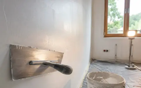 Nakładanie gładzi na ścianę za pomocą kielni. W tle okno i lampa budowlana.