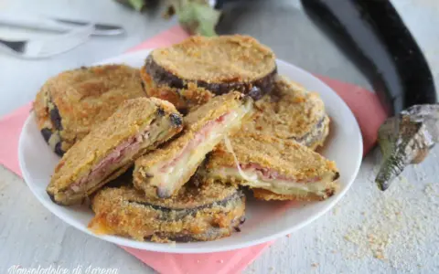 Cordon bleu di melanzane impanati e fritti, ripieni di prosciutto e formaggio filante, serviti su un piatto bianco con melanzane fresche sullo sfondo.