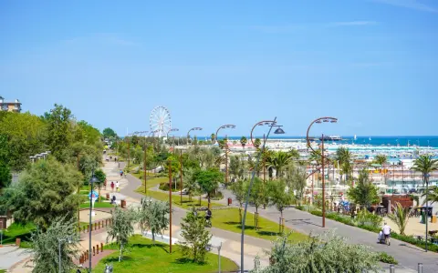 Passeggiata nel verde del parco del mare Rimini, con gente che cammina e pedala, e la ruota panoramica sullo sfondo.