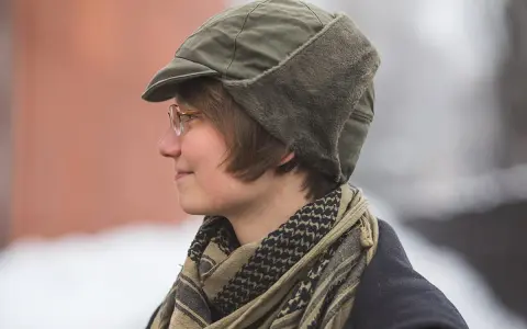 Osoba w okularach nosi zieloną czapkę polową z futrzanymi nausznikami i szalik w stylu keffiyeh.