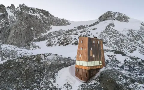 Il rifugio Sasso Nero, un'architettura moderna in legno, si erge solitario tra rocce e neve, dominato da imponenti vette montuose.