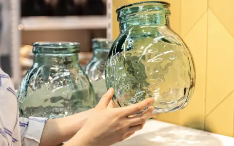 Une main examine un objet en verre texturé, vert pâle, aux reflets ondulés. D'autres objets similaires sont visibles en arrière-plan.