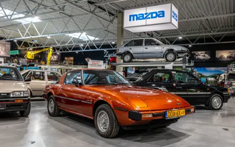 Pomarańczowa Mazda RX-7 błyszczy w muzeum samochodów, obok innych klasyków marki.
