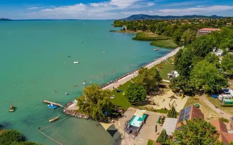 Słoneczny dzień na kempingu Balaton. Ludzie kąpią się w jeziorze i basenie, bawią na placu zabaw, a kampery stoją wśród drzew.