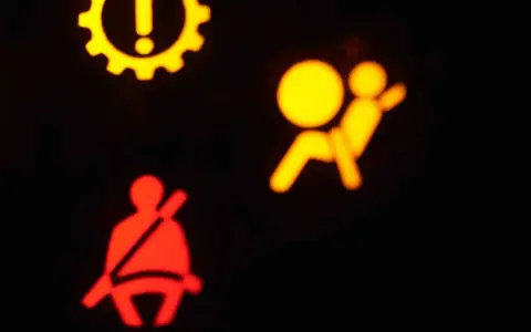 Das gelbe airbag symbol leuchtet. Daneben ein rotes Symbol für den Sicherheitsgurt. Ein gelbes Zahnrad mit Ausrufezeichen ist oben links zu sehen.