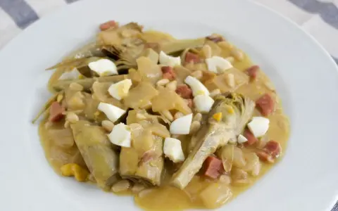 Plato de alcachofas con jamón, huevo duro y piñones, servido en una cazuela.