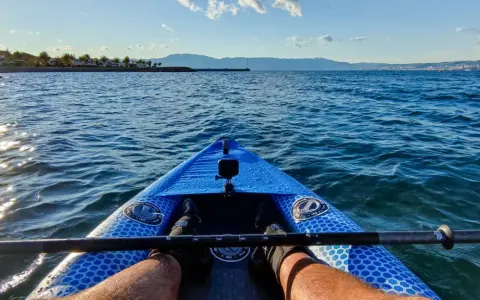 Płynę niebieską kanadyjką po spokojnym morzu. Słońce odbija się w wodzie, a w oddali widać góry i miasto.