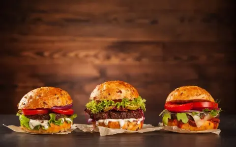 Trzy apetyczne burgery z różnymi dodatkami na drewnianym tle.