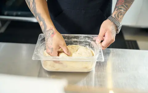 Ręce z tatuażami zagniatają ciasto w plastikowym pojemniku. To pierwszy krok, by dowiedzieć się, jak mrozić ciasto.