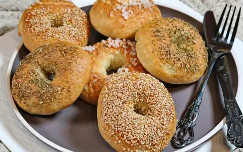 Sechs selbstgemachte Bagels auf einem Teller: zwei mit Sesam, zwei mit Kräutern und zwei mit grobem Salz bestreut. Ein Messer und eine Gabel liegen daneben.
