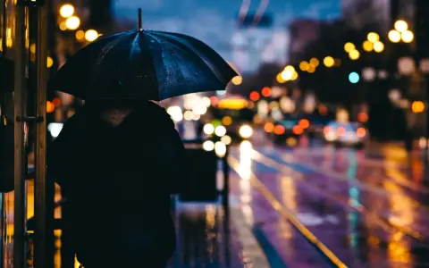 Person sous un parapluie noir, attendant sur un trottoir mouillé. Les lumières de la ville se reflètent sur la chaussée, créant de belles photos de pluie.