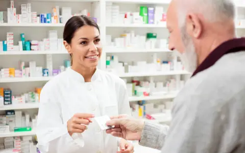 Uśmiechnięta farmaceutka podaje starszemu panu leki. W tle apteczne półki pełne leków, w tym poltixa.
