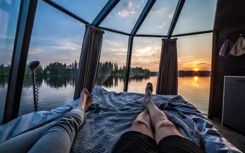 Två personer vilar i en glaskupa med utsikt över en sjö i Norrland vid solnedgången. En perfekt glampingupplevelse.