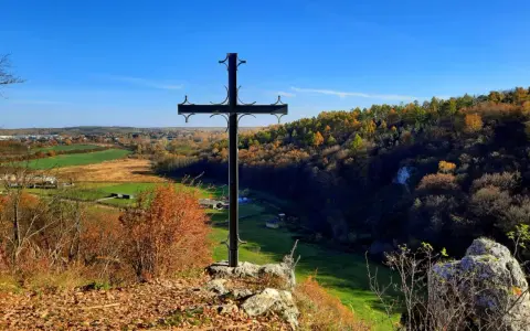 Czarny krzyż na skale góruje nad jesienną doliną Grzybowską. W oddali widać pola i lasy w barwach złota i czerwieni.