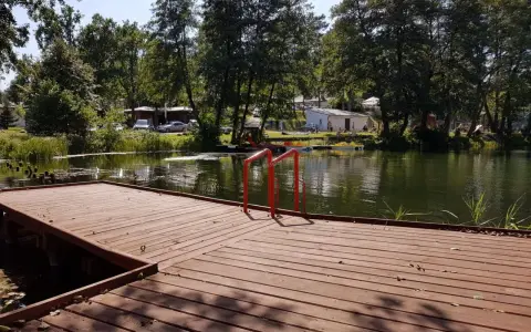 Drewniany pomost z czerwonymi poręczami nad jeziorem. W tle drzewa, budynki i samochody. Idealne miejsce na łagów camping.