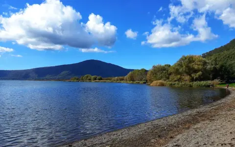 Spiaggia ghiaiosa e acque calme del lago di Vico, con montagne verdi e cielo azzurro punteggiato di nuvole bianche.