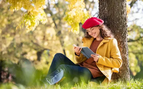 Mujer con boina roja lee un libro bajo un árbol, inspirada por los temas de cuentos del otoño.