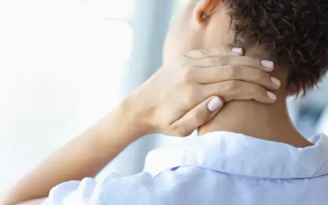 Frau mit Hand am Nacken, die Schmerzen andeutet. Wann zum Arzt bei Nackenschmerzen?