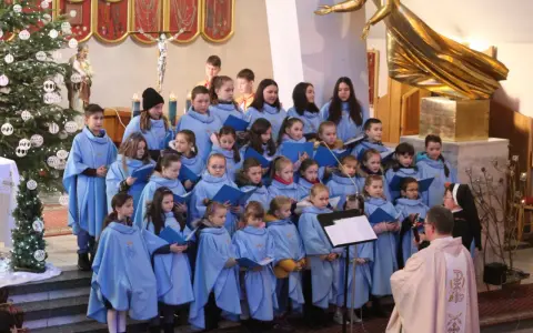 Dzieci w błękitnych strojach tworzą schola, śpiewając w kościele przy choince.