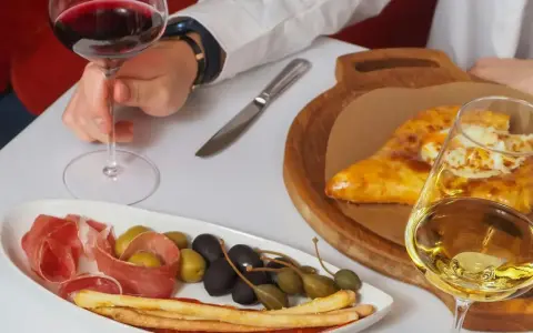 Ręka trzyma kieliszek czerwonego wina wytrawnego. Obok talerz z wędliną, oliwkami i grissini oraz chaczapuri i kieliszek białego wina.