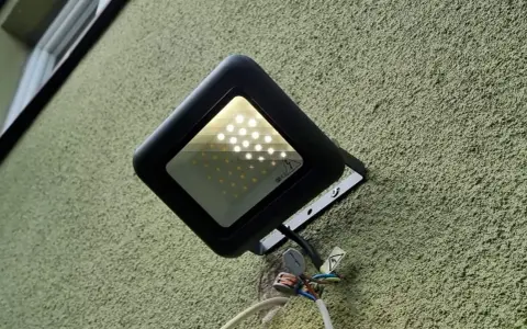 Czarna lampa led żarzy się po włączeniu, oświetlając zieloną, chropowatą ścianę. Widać przewody i złączki.