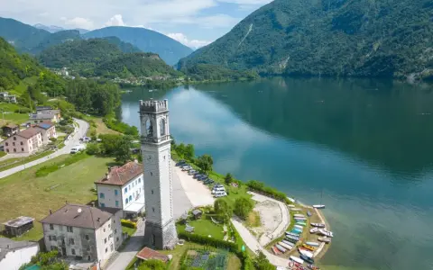 Torre campanaria affacciata sul lago di Corlo, con montagne verdi sullo sfondo e un villaggio pittoresco.