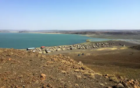 Widok na osadę nad turkusowym jeziorem Turkana. W oddali widać górzysty krajobraz.