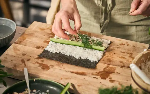 Händer lägger dill på hemmagjord sushi med ris, gurka och fyllning på en skärbräda.