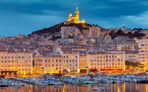 Porto di Marsiglia illuminato al tramonto, con la Basilica di Notre-Dame de la Garde in cima alla collina. Perfetto per le tue marsiglia escursioni fai da te.