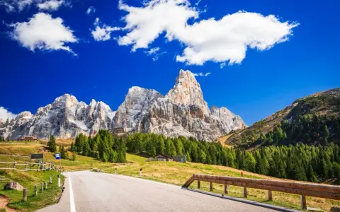 Strada panoramica per esplorare i passi del Trentino in moto, con imponenti vette dolomitiche sullo sfondo e un cielo azzurro con nuvole bianche.