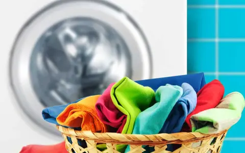 Cesto pieno di panni colorati pronti per scoprire come lavare i colorati in lavatrice. Lavatrice sullo sfondo.