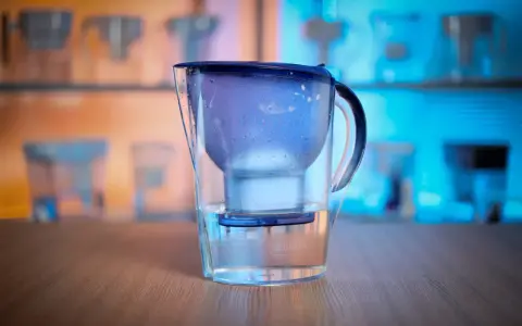 En vattenrenare för dricksvatten, en Brita-kanna, står på ett träbord. Bakgrunden visar fler liknande kannor i blått och orange ljus.