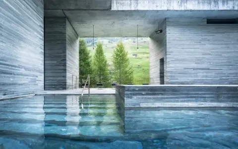 Piscine intérieure des thermes de Vals Zumthor, avec vue sur les montagnes verdoyantes et les sapins. L'eau bleue reflète les murs en pierre grise.
