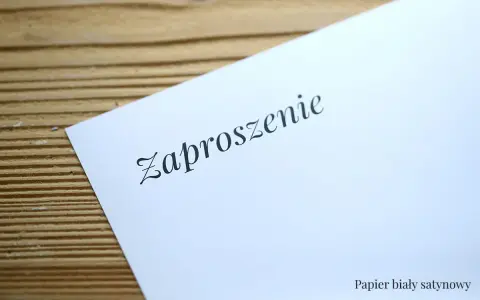 Eleganckie zaproszenie na ślub na białym, satynowym papierze, leżące na drewnianym tle.