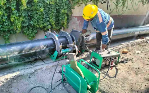 Un ouvrier utilise une machine de soudure pour joindre un gros tuyau noir. L'équipement comprend une douille à souder et une unité de contrôle verte.