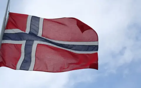 Flaga Norwegii powiewa na wietrze na tle błękitnego nieba z chmurami.