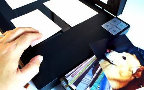 Ręka kładzie zdjęcie na skanerze, rozpoczynając proces digitalizacji zdjęć. Obok stos innych fotografii.