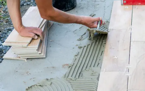 Układanie płytek na betonowym tarasie. Ręce pracownika rozprowadzają klej, obok stos płytek i poziomica.