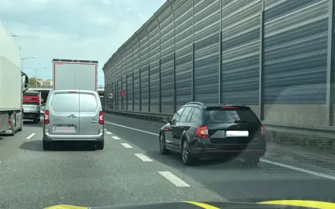 Korek na autostradzie. Samochody osobowe i ciężarowe stoją w rzędzie. W tle widać ekrany akustyczne. Jazda na suwak przepis to podstawa.