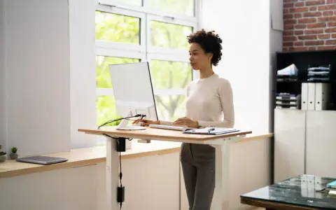 Une femme travaille sur un ordinateur à un bureau réglable en hauteur. La position debout prolongée présente des risques pour la santé, il est donc important de varier les postures.