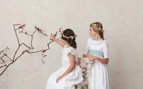 Dos niñas con vestidos blancos y coronas de flores, una sentada tocando una rama, la otra de pie. Peinados de trenza para niña de comunión.