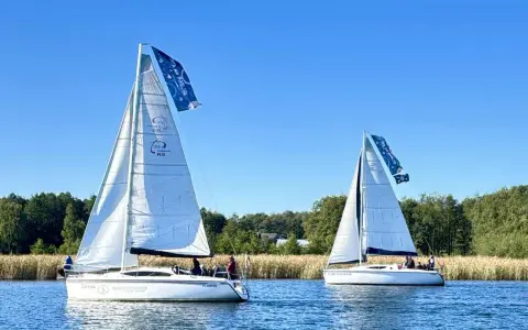 Dwa żaglowce na jeziorze, symbolizujące to, z czego słyną Mazury – żeglarstwo i piękne krajobrazy.