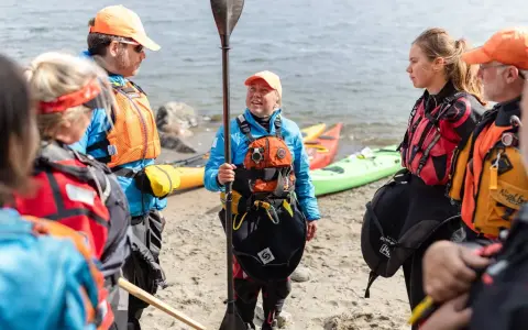 Grupp samlad på stranden, redo för paddling. En instruktör visar paddelteknik. Kajaker ligger i bakgrunden.