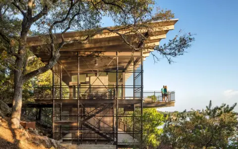 Maison moderne sur un terrain en pente raide, avec des balcons et des escaliers extérieurs en métal, nichée dans les arbres près d'un lac.