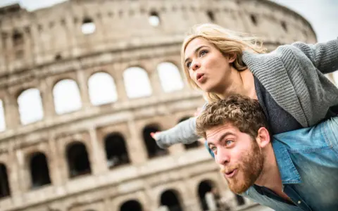 Coppia in una vacanza romantica a Roma, con il Colosseo sullo sfondo. Lui porta lei sulle spalle, entrambi con espressioni sorprese.