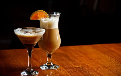 Dwa drinki z likierem kawowym i mlekiem: jeden w kieliszku martini, drugi w wysokiej szklance z pomarańczą.